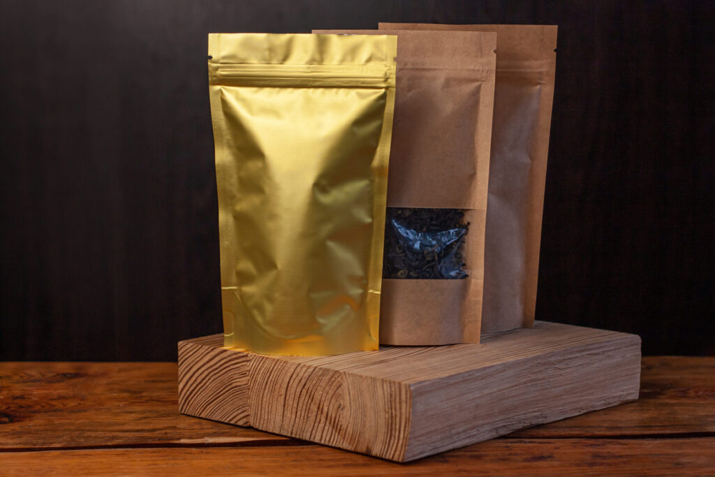 Brown kraft paper pouch bags with coffee beans and tea leaves in wicker basket with white textile on wooden background.